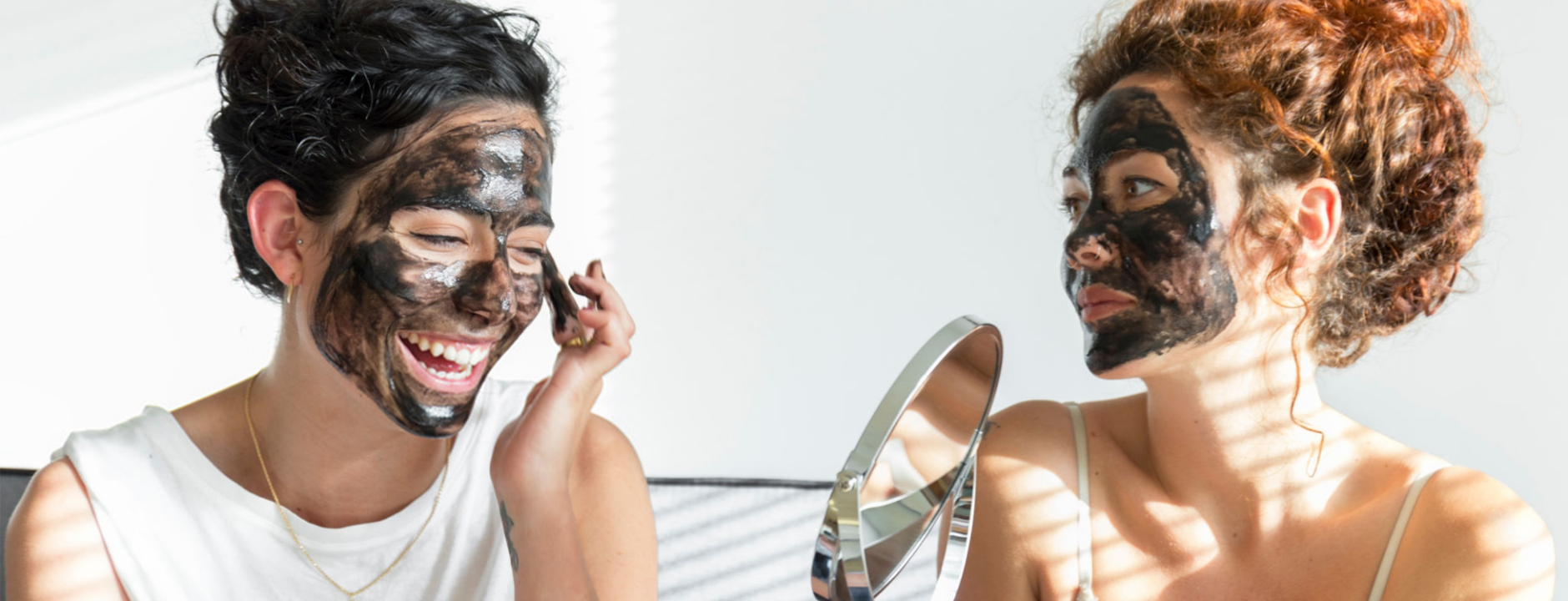 Two people applying black face masks, one looking into a mirror.